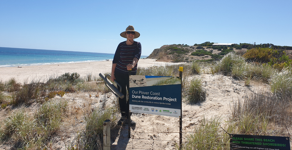 Person standing next to an 'Our Plover Coastal Dune Restoration Project' sign on a beach.