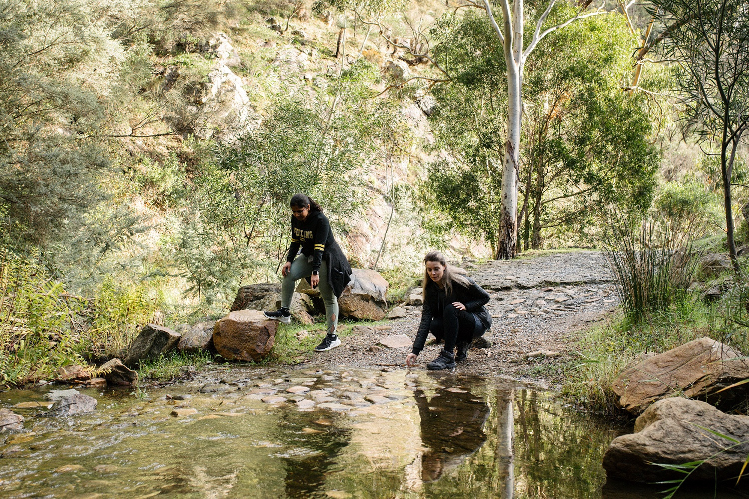 Two people standing at the edge of a waterway in Adelaide, one crouched looking at the water.