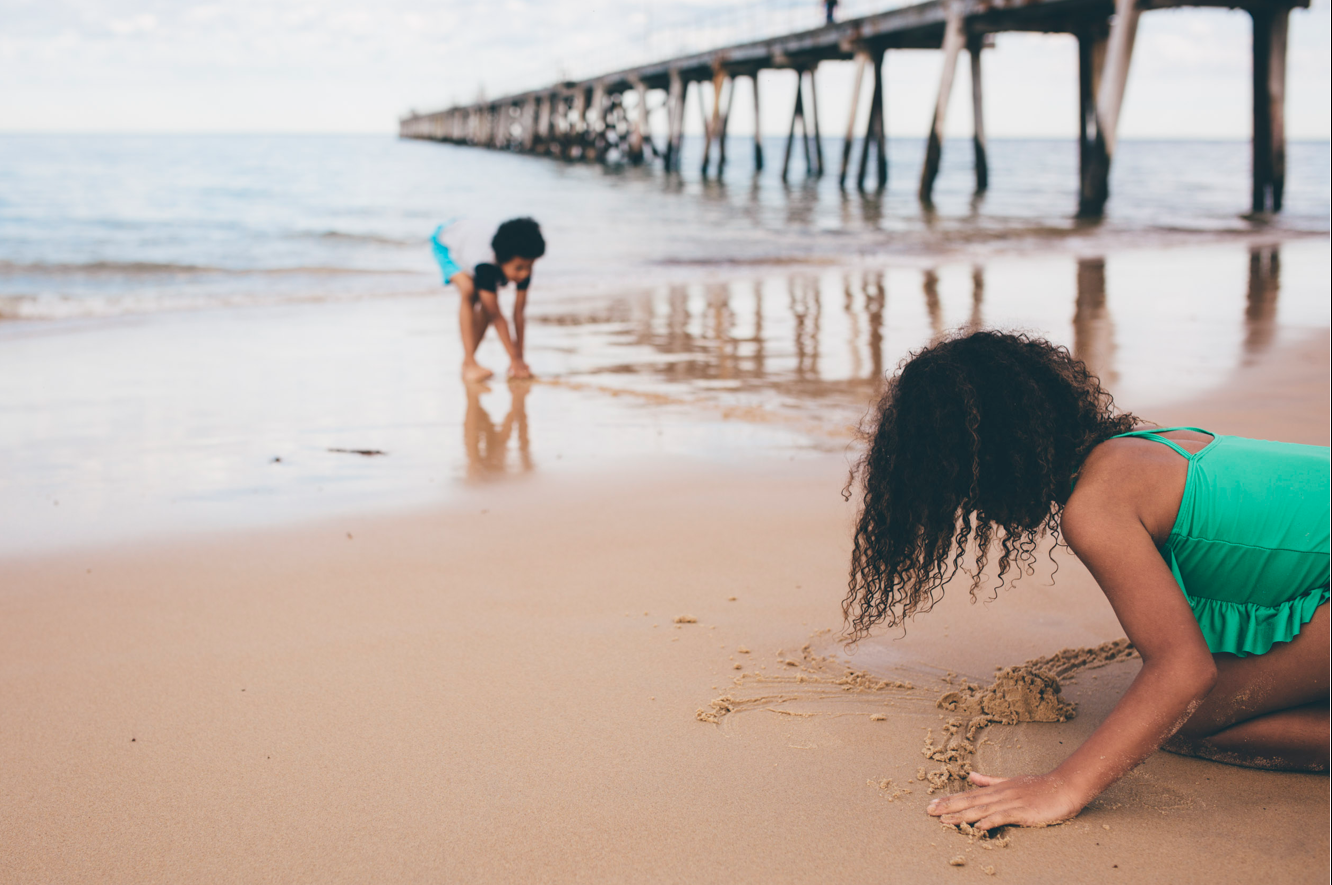 Two kids play on a beach near a jetty, one close to camera wears green bathers, and one further away has a blue shovel