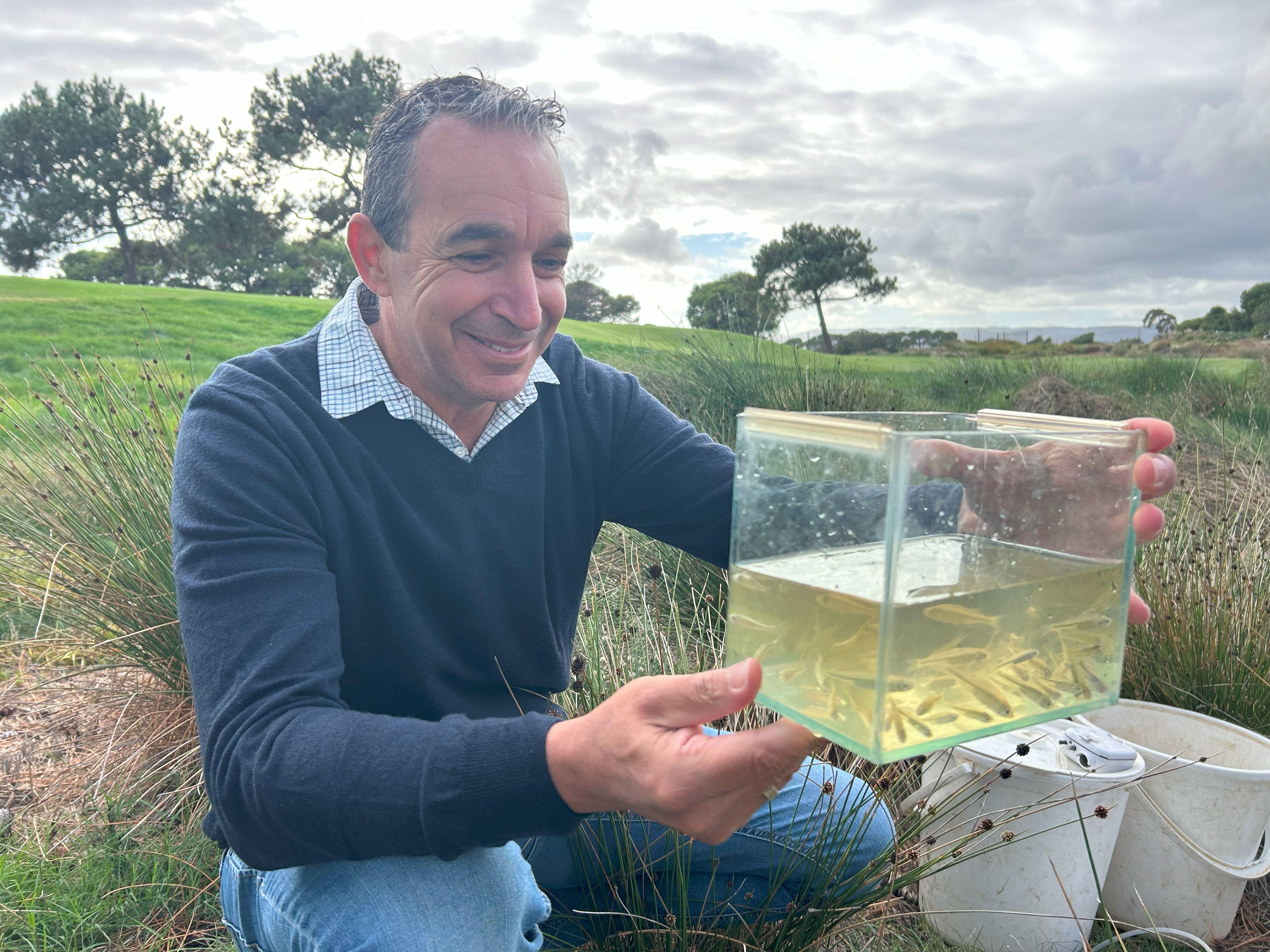 A man crouches holding a clear container with small fish