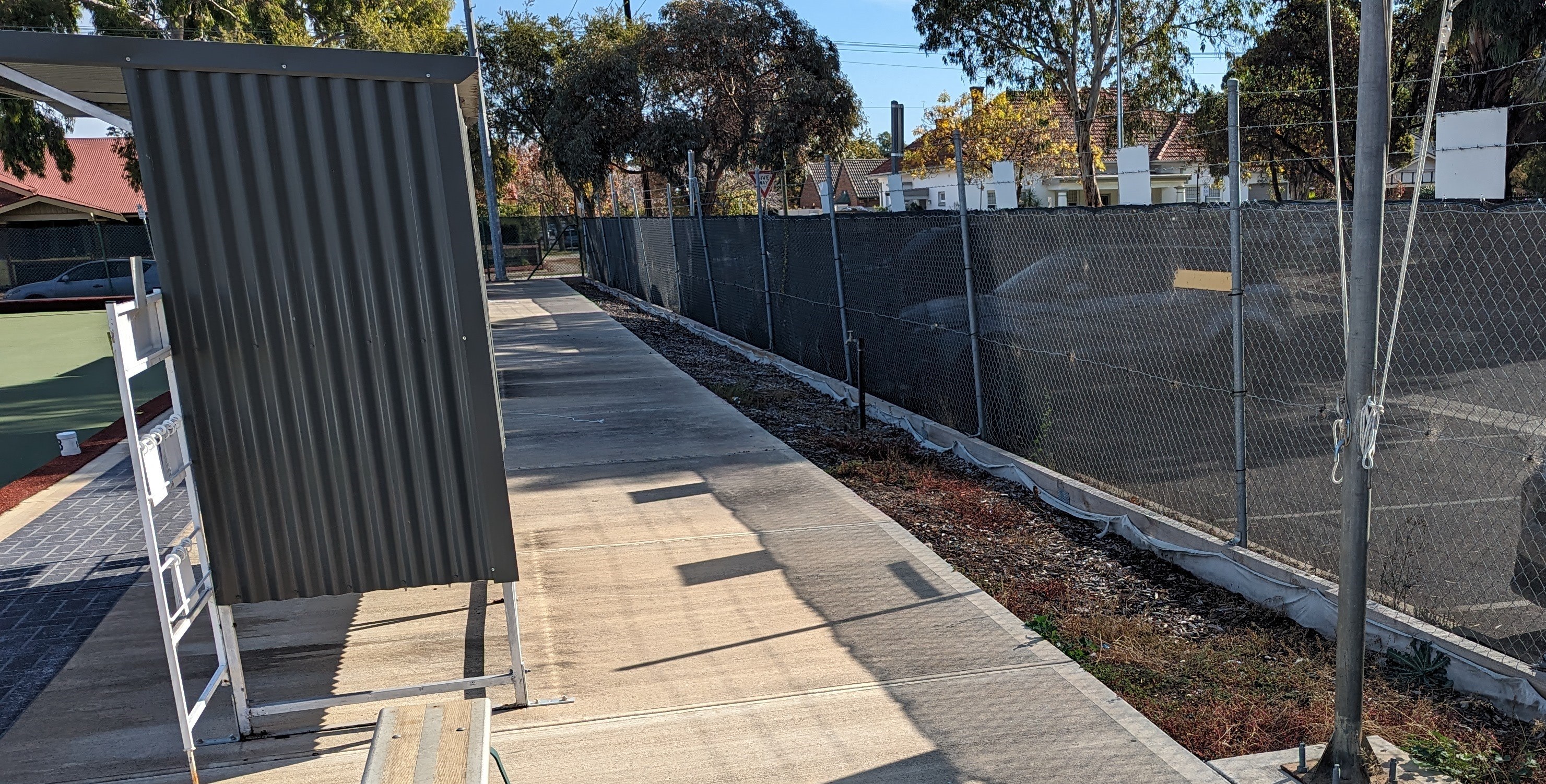 Woodville Bowling Club's shelter, path and car park divided by a wire fence. There's a strip of dirt along the fence line where plants will be placed.