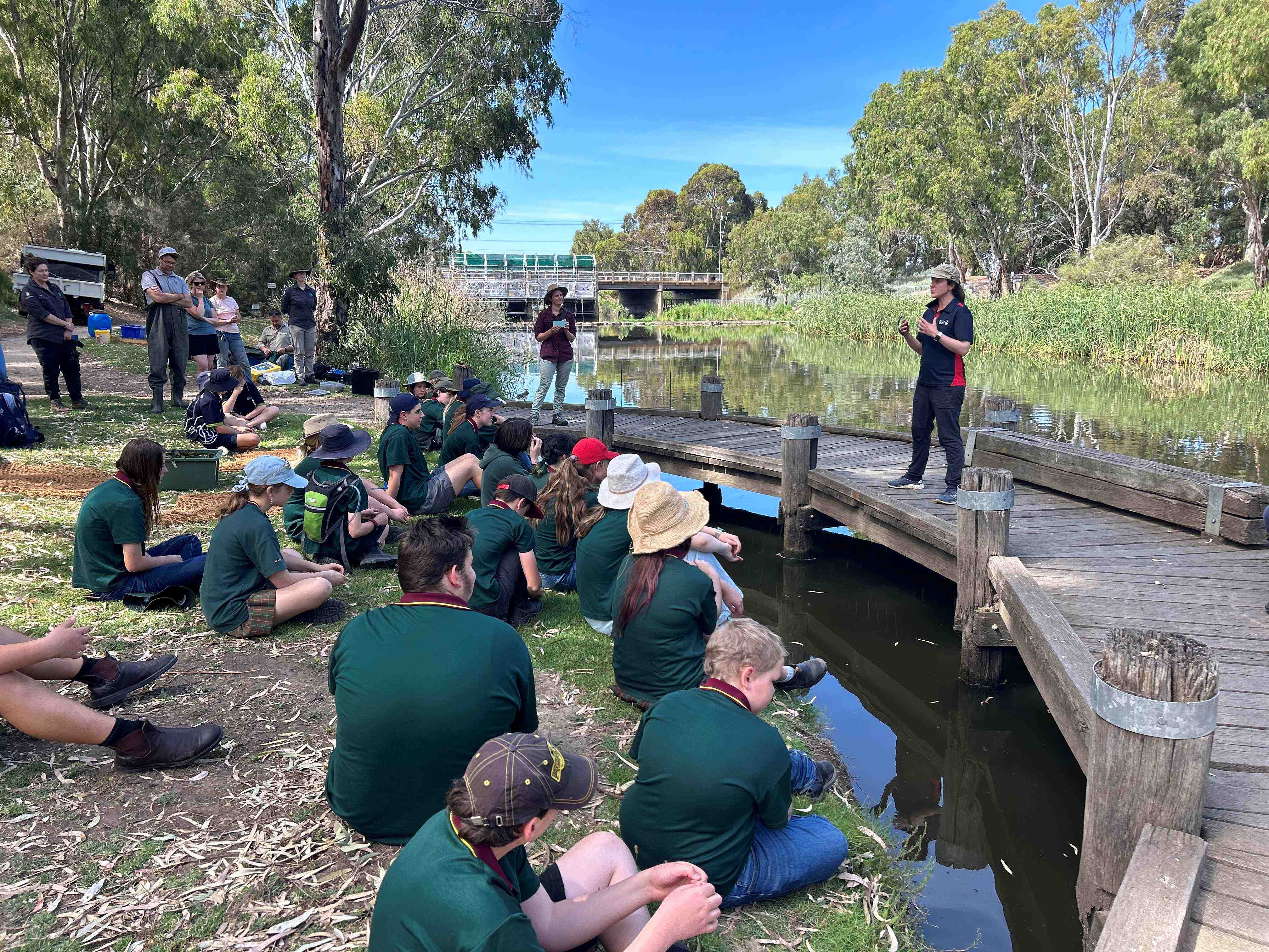 A woman talking to students sitting on a bank by a river.